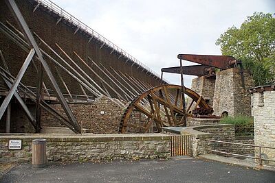 Auf dem Bild ist ein Wasserrad mit  Gradierwerk im Hintergrund zu sehen.