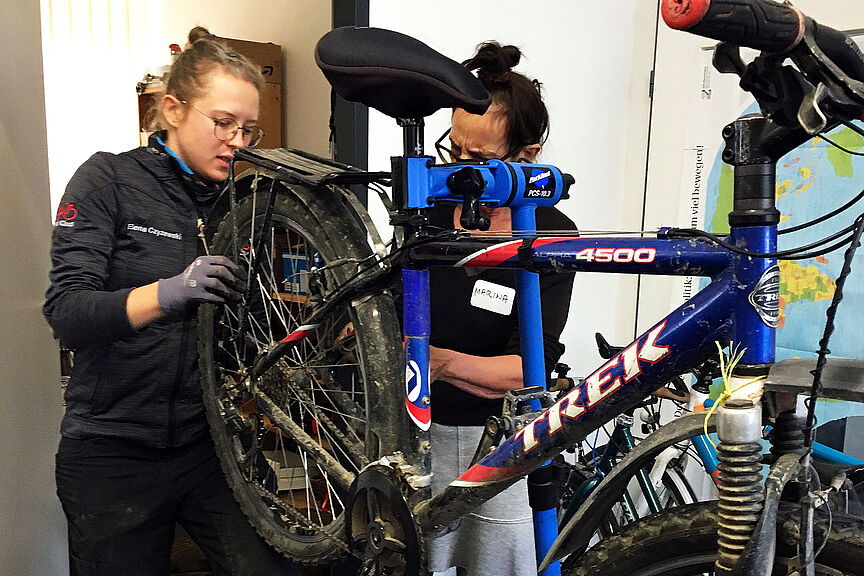 Fahrrad-Schrauberinnenkurs des ADFC Bad Naueim Friedberg Zwei Frauen hantieren an einem aufgeständerten Fahrrad und scheinen etwas zu reparieren.