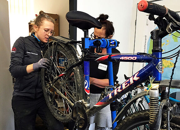 Zwei Frauen hantieren an einem aufgeständerten Fahrrad und scheinen etwas zu reparieren.