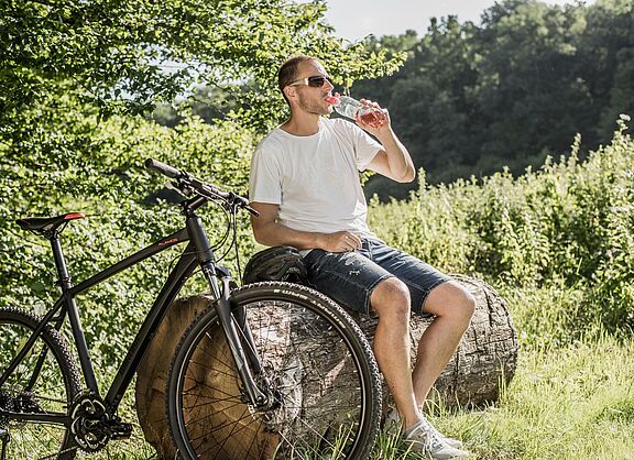 Radfahrer mit Sonnenbrille trinkt Wasser aus einer Flasche während einer Pause auf einem Baumstamm. Sein schwarzes Mountainbike steht daneben.