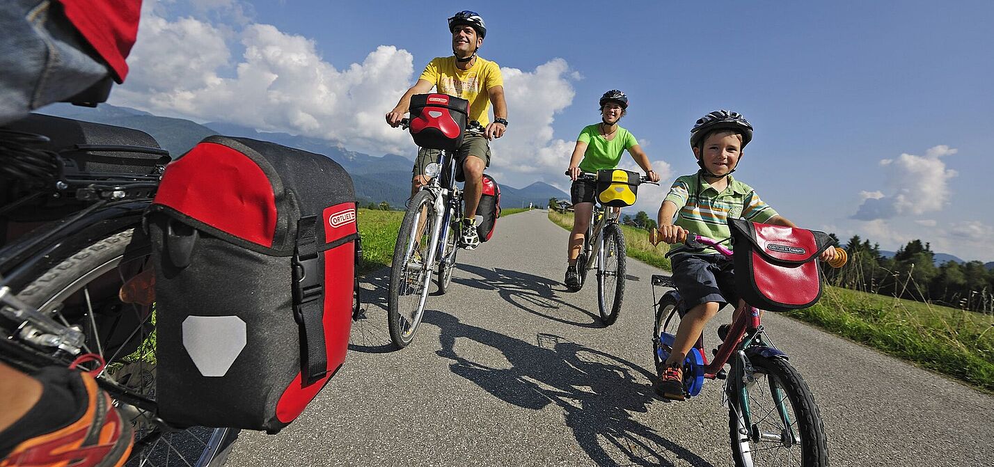 Eine vierköpfige Familie fährt mit Helmen und Packtaschen auf Fahrrädern eine schmale Landstraße entlang; vorn ein Kind auf einem kleinen Rad, dahinter zwei Erwachsene und ein weiteres Fahrrad im Anschnitt links, umgeben von Wiesen, Bergen und blauem Himmel.